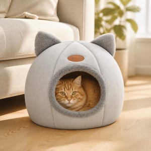 May include: A light gray, dome-shaped pet bed with two plush, dark gray cat ears on top. The bed features a circular opening with a textured gray trim and a small, oval-shaped tan patch on the front. An orange tabby cat is resting inside the bed.