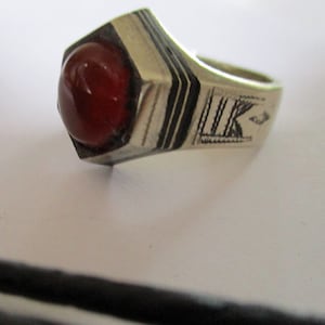 May include: A silver ring with a red carnelian stone set in a hexagonal bezel. The band is decorated with black enamel and intricate engravings.