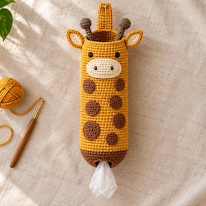 May include: A yellow and brown crocheted giraffe-shaped plastic bag dispenser with a hanging loop at the top, dark brown spots, a cream-colored muzzle, and a small opening at the bottom where a white plastic bag is visible.