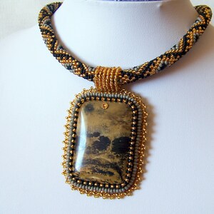 Pu&ograve; includere: Una collana di perline con uno schema di colori oro, nero e grigio. La collana ha un grande ciondolo rettangolare con una pietra marrone e nera.