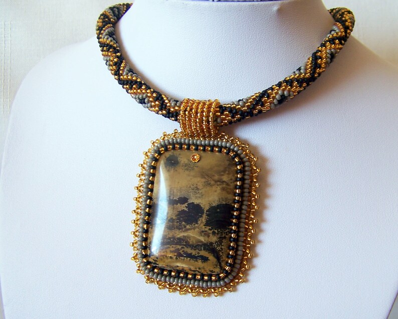 Pu&ograve; includere: Una collana di perline con uno schema di colori oro, nero e grigio. La collana ha un grande ciondolo rettangolare con una pietra marrone e nera.