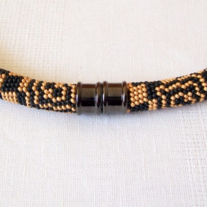 May include: A black and gold beaded necklace with a magnetic clasp. The necklace is made with small, round beads in a geometric pattern.