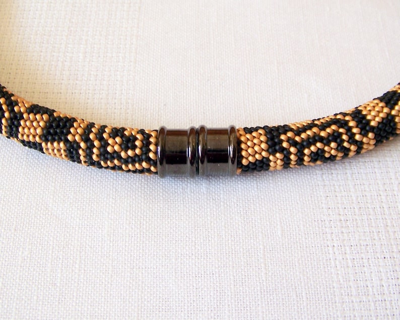 May include: A black and gold beaded necklace with a magnetic clasp. The necklace is made with small, round beads in a geometric pattern.