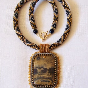 Pu&ograve; includere: Una collana di perline con uno schema di colori nero, oro e grigio. La collana ha un ciondolo rettangolare con una pietra marrone e nera.