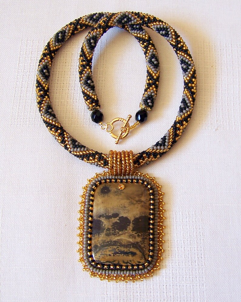 Pu&ograve; includere: Una collana di perline con uno schema di colori nero, oro e grigio. La collana ha un ciondolo rettangolare con una pietra marrone e nera.