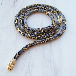 Könnte beinhalten: Eine Perlenkette in Schlangenform mit einem goldenen und grauen Muster. Die Kette hat einen goldenen Schlangenkopf-Anhänger.