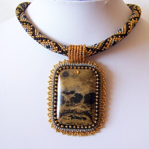 Pu&ograve; includere: Una collana di perline con uno schema di colori oro, nero e grigio. La collana presenta un grande ciondolo rettangolare con una pietra marrone e nera.