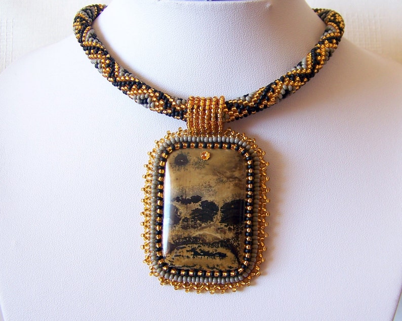 Pu&ograve; includere: Una collana di perline con uno schema di colori oro, nero e grigio. La collana presenta un grande ciondolo rettangolare con una pietra marrone e nera.