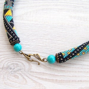 May include: A beaded necklace with a geometric pattern in black, gray, turquoise, and yellow. The necklace has a turquoise bead and a bronze clasp.