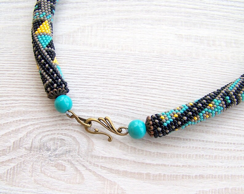 May include: A beaded necklace with a geometric pattern in black, gray, turquoise, and yellow. The necklace has a turquoise bead and a bronze clasp.