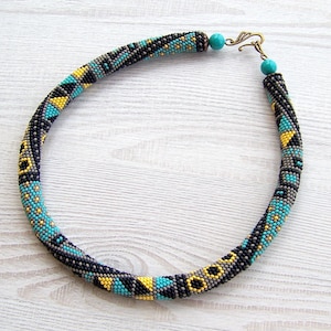 May include: A beaded necklace with a geometric pattern in shades of black, gray, turquoise, and yellow. The necklace has a simple clasp.