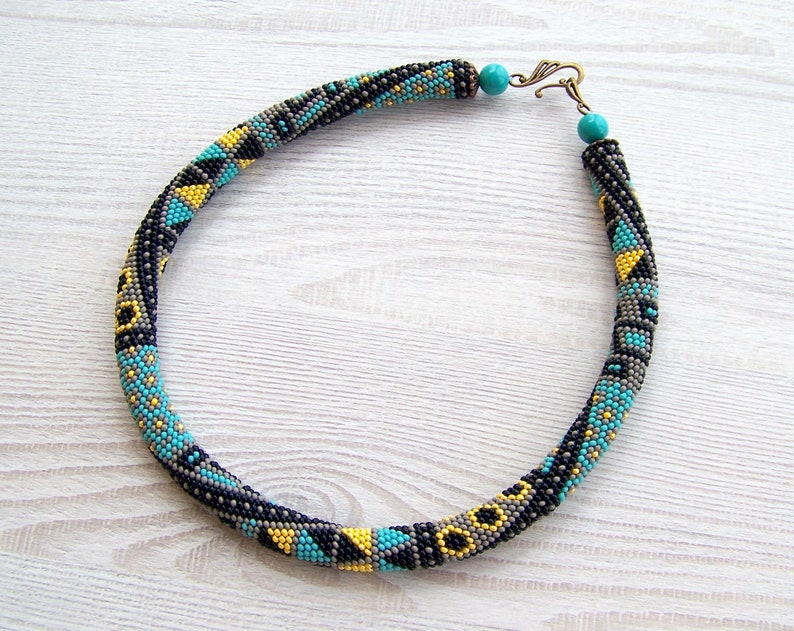 May include: A beaded necklace with a geometric pattern in shades of black, gray, turquoise, and yellow. The necklace has a simple clasp.