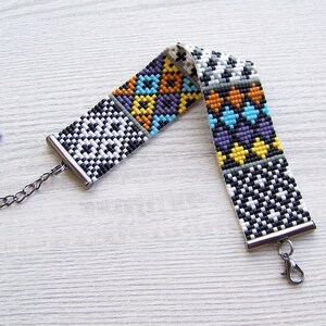 May include: A colorful beaded bracelet with a geometric pattern in black, white, yellow, orange, blue, and purple. The bracelet has a silver clasp and chain.