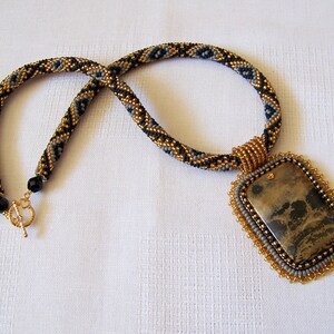 Pu&ograve; includere: Una collana di perline con un motivo geometrico dorato e nero e un grande ciondolo rettangolare con una pietra marrone e nera.