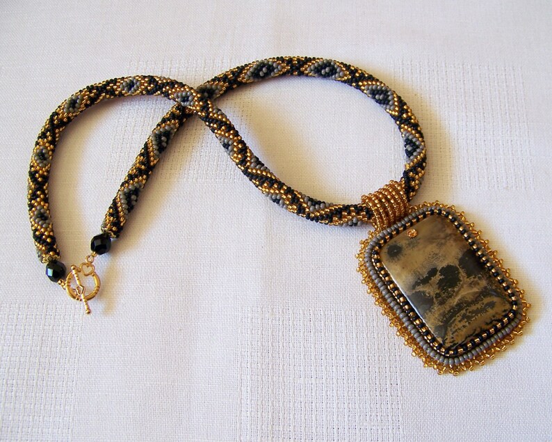 Pu&ograve; includere: Una collana di perline con un motivo geometrico dorato e nero e un grande ciondolo rettangolare con una pietra marrone e nera.