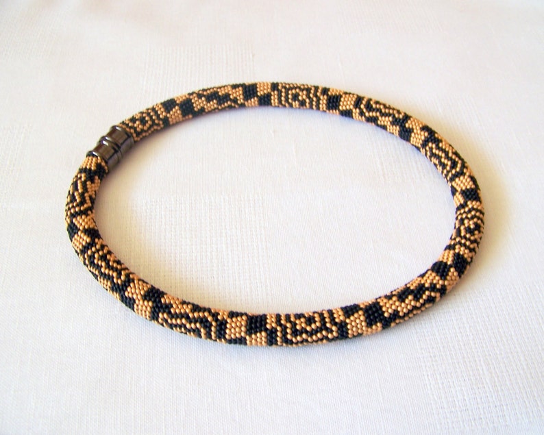 May include: A black and gold beaded necklace with a magnetic clasp. The necklace is made of small, round beads that are arranged in a geometric pattern.