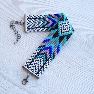 Puede incluir: Un brazalete de cuentas con un patr&oacute;n geom&eacute;trico en tonos de azul, negro, blanco y rosa. El brazalete tiene un cierre y una cadena plateados.