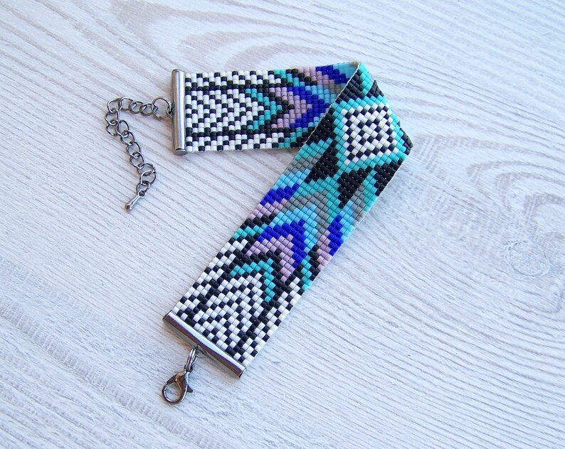 Puede incluir: Un brazalete de cuentas con un patr&oacute;n geom&eacute;trico en tonos de azul, negro, blanco y rosa. El brazalete tiene un cierre y una cadena plateados.