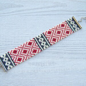 May include: A white, red, and black beaded bracelet with a geometric pattern. The bracelet has a silver clasp and extender chain.