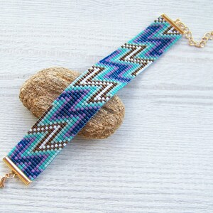 May include: A beaded bracelet with a geometric pattern in shades of blue, purple, white, and brown. The bracelet has a gold clasp and extender chain.