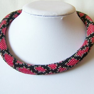 以下が含まれることがあります： 幾何学模様の赤い、黒い、銀色のビーズネックレス。ネックレスは白いマネキンに乗っています。