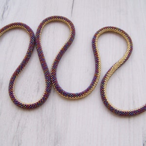 May include: A gold and purple beaded necklace with a snake-like design. The necklace is made of small, round beads that are woven together in a tight pattern.