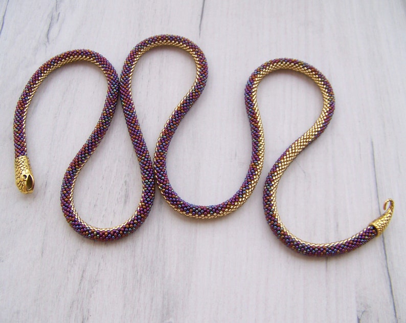 May include: A gold and purple beaded necklace with a snake-like design. The necklace is made of small, round beads that are woven together in a tight pattern.