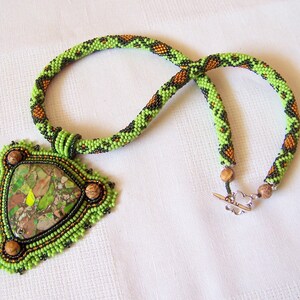 May include: A green, black, and brown beaded necklace with a triangular pendant featuring a green and brown stone. The pendant is surrounded by smaller beads in green, black, and brown.