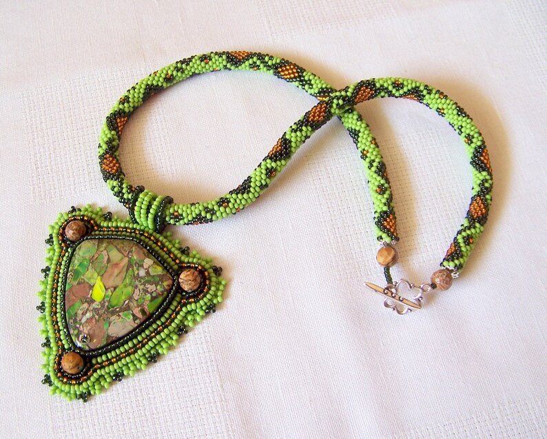 May include: A green, black, and brown beaded necklace with a triangular pendant featuring a green and brown stone. The pendant is surrounded by smaller beads in green, black, and brown.