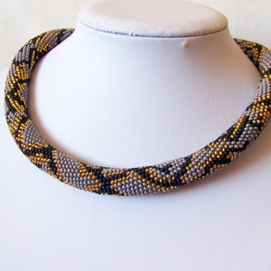 Può includere: Una collana di perline grigie, nere e dorate con un motivo a pelle di serpente.