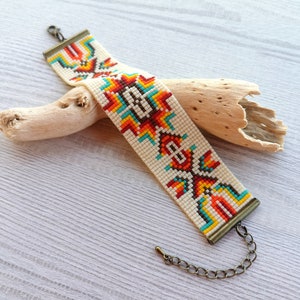 May include: A white beaded bracelet with a colourful geometric pattern in red, orange, yellow, blue, and turquoise. The bracelet has a metal clasp and chain.