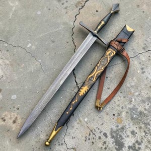 May include: A double-edged steel sword with a pointed tip, a crossguard, and a dark handle. Beside it lies a black scabbard featuring gold-colored decorative filigree, a metal tip, and a brown leather strap for carrying the weapon.