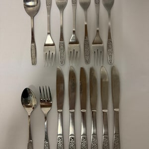 May include: A set of stainless steel flatware consisting of two spoons, six forks, and six knives. Each piece features a polished finish and a decorative floral pattern embossed on the handle. The utensils are arranged in two rows on a plain surface.