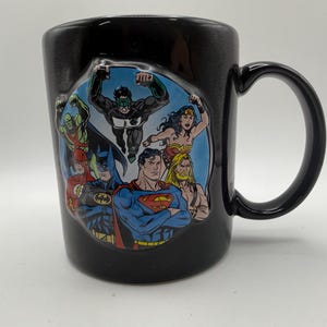 Peut inclure: Un mug en céramique noire orné d'un graphique circulaire représentant des super-héros de DC Comics, dont Superman, Batman, Wonder Woman, Green Lantern, Flash, Aquaman et Martian Manhunter, illustrés dans un style de bande dessinée classique.