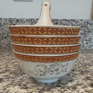 May include: A stack of three white ceramic bowls with a central vertical handle. Each bowl features a wide band of orange with a repeating yellow geometric pattern and a gold rim. The base of the bottom bowl displays a delicate gold floral design.