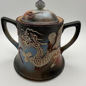 May include: A ceramic lidded sugar bowl featuring a raised relief dragon design in cream and brown tones. The body has a gradient finish transitioning from dark brown to light blue, accented with white beaded detailing along the edges and handles.