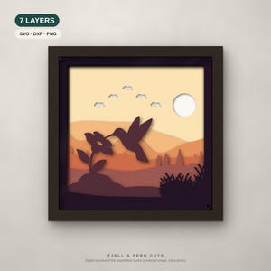 Op de afbeelding: Een vierkant 7-laags papierkunstwerk in een donkere lijst met een kolibrie boven een bloem. De scène bevat een zon, wolken en gelaagde heuvels in geel-, oranje- en paarstinten. Tekst: 7 LAYERS, SVG, DXF, PNG en FJELL & FERN CUTS.