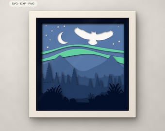 Op de afbeelding: Een vierkant ingelijst 7-laags papierkunstwerk met een nachtlandschap met een witte uil, een maansikkel, sterren en groen noorderlicht boven blauwe bergen en dennenbomen. Tekst: 7 LAYERS, SVG, DXF, PNG, FJELL & FERN CUTS.