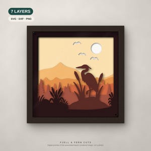 May include: A square 7-layer paper cut art piece in a dark frame featuring a heron silhouette in a marshland landscape with mountains, cattails, and birds under a sun. Text labels include 7 LAYERS, SVG, DXF, PNG, and FJELL & FERN CUTS.