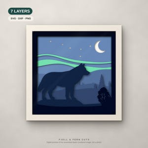 Op de afbeelding: Een zevenlaags papierknipkunstwerk in een vierkante lijst met het silhouet van een donkerblauwe wolf voor een landschap met lichtgroen noorderlicht, een witte maansikkel en sterren tegen een blauwe lucht. Tekst: 7 LAYERS.