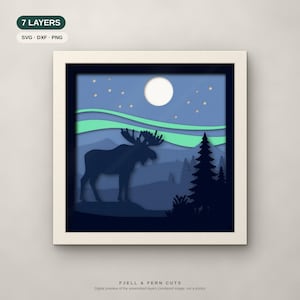 Op de afbeelding: Een vierkant ingelijst 7-laags papierkunstwerk met een donkerblauw eland-silhouet in een boslandschap onder een lichtblauwe lucht met een witte maan, sterren en groen noorderlicht. Tekst bevat 7 LAYERS, SVG, DXF, PNG en FJELL & FERN CUTS.