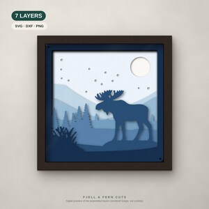 Op de afbeelding: Een vierkant ingelijst meerlagig papierkunstwerk met een donkerblauw eland-silhouet in een boslandschap met bergen, een maan en sterren. De tekst 7 LAYERS, SVG, DXF, PNG en FJELL & FERN CUTS is zichtbaar.