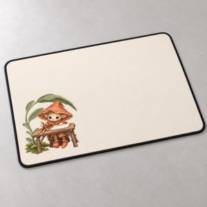 Puede incluir: Una alfombrilla de escritorio rectangular con superficie color crema y borde negro cosido. En la esquina inferior izquierda, una pequeña criatura con sombrero naranja está sentada en un escritorio de madera escribiendo en un papel.