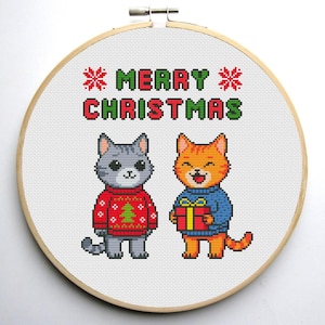 May include: Cross-stitch hoop art featuring two cartoon cats in sweaters. One gray cat wears a red sweater with a Christmas tree, the other orange cat holds a gift. The text "MERRY CHRISTMAS" is stitched above the cats.