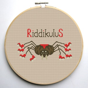 May include: Cross-stitch embroidery hoop featuring a brown spider with red eyes and red legs, surrounded by red and white mushrooms. The text "Riddikulus" is stitched above the spider.