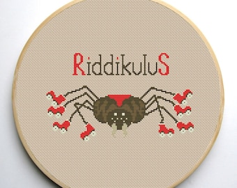 May include: Cross-stitch embroidery hoop with a brown spider with red eyes and red legs, surrounded by red and white mushrooms. The text "Riddikulus" is stitched above the spider.