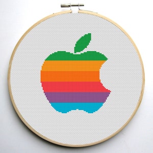May include: A rainbow-colored Apple logo cross-stitched on white fabric in a wooden embroidery hoop.
