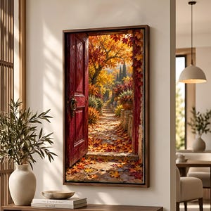 May include: A rectangular wall art print in a dark wood frame depicting an open red door revealing a stone path covered in fallen autumn leaves. The scene features vibrant orange and yellow trees, stone walls, and foliage under a bright sky.