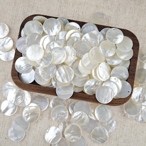 May include: A collection of round, flat, white mother-of-pearl shell buttons, each featuring two small holes for sewing. The buttons are scattered both inside and around a rectangular wooden tray, displaying a natural iridescent sheen.
