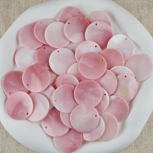 May include: A collection of circular, flat, light pink shell beads, each featuring a single small hole near the edge for threading. The beads have a smooth, polished surface with subtle, swirling pearlescent variations in shades of soft pink and white.